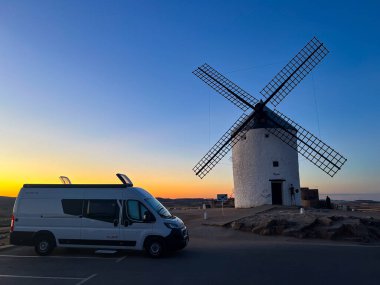 Consuegra, İspanya; 23 Eylül 2025: Kamp minibüsü renkli bir günbatımı gökyüzünün altında geleneksel bir İspanyol yel değirmeninin yanına park edildi