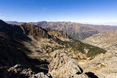 Bir vadi, kayalık tepeler ve mavi gökyüzünün altında dağınık bitki örtüsü barındıran engebeli dağlık bir arazi.