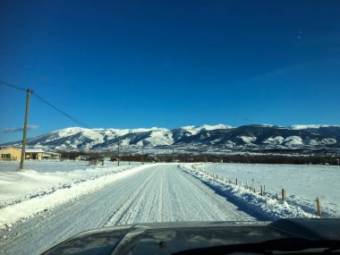 Soğuk bir kış gününde, açık mavi gökyüzünün altında, uzak dağlara giden yolu karla kaplayan yol.