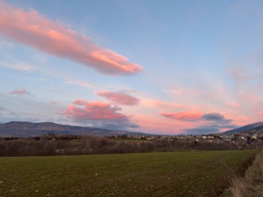 Pembe merceksi bulutlar yeşil bir tarla ve köy üzerinde süzülür, yumuşak akşam ışığı ile yıkanır.