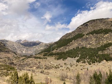 Dağ vadisi manzarasında bulutlu bir gökyüzünün altında hidroelektrik baraj, sütunlar ve dağınık çam ağaçları bulunur.