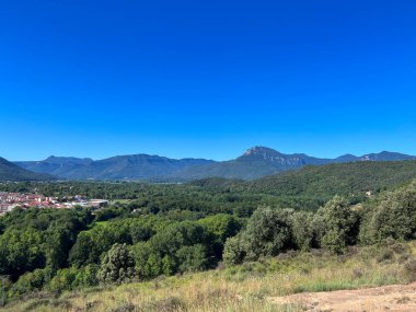 Panoramik manzara Olot şehrini, yeşil ağaçları ve açık gökyüzünün altındaki uzak dağları yakalıyor.