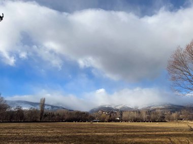 Yuvarlanan tarlalar ve çıplak ağaçlar bulutlu bir gökyüzünün altında kar kaplı dağları olan bir köye gidiyor.