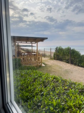 Pencere manzaralı plaj, deniz, açık hava restoranı ve yeşil çalılar.