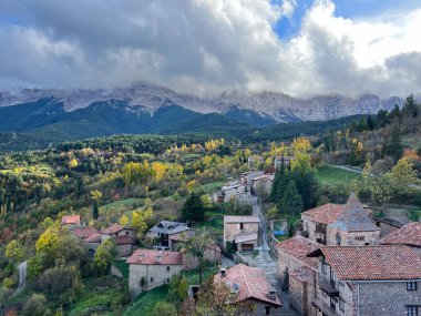 Kırsal Pireneler köyü, rengarenk ağaçlar ve bulutlu bir gökyüzünün altındaki kayalık dağlarla dolu bir sonbahar manzarasına yuva yapmış.