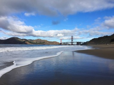 Okyanus dalgaları tarafından çerçevelenmiş Golden Gate Köprüsü kumsalda kırılıyor ve tepeler uzaklaşıyor.
