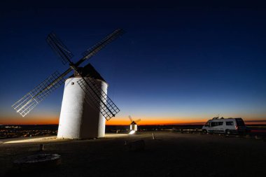 Beyaz yel değirmenleri, renkli bir alacakaranlık gökyüzünün altında park etmiş bir karavanla tepenin üstünde duruyor.