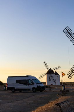Gün batımında tarihi yel değirmenlerinin yanına park edilmiş bir karavan ve bir İspanyol bayrağı.