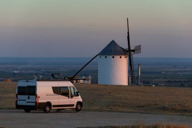 Campo de Criptana 'da günbatımında tarihi yel değirmenine hayran olan bir kamp aracı tepede park etmiş.