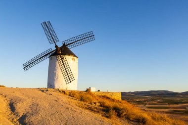 Consuegra yel değirmeni altın saat ışığıyla aydınlanan bir tepenin üzerinde duruyor, açık bir gökyüzünün altında.