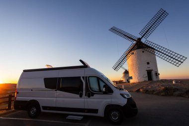 Altın saatinde tarihi yel değirmenlerinin yakınına park etmiş bir karavan, seyahat macerası ve özgürlüğü resmediyor.