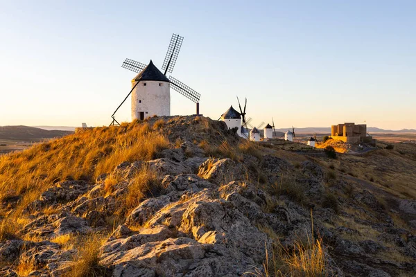 Tarihsel yel değirmenleri ve sıcak günbatımı ışığıyla aydınlatılmış bir tepenin üzerinde duran bir ortaçağ şatosu.