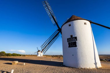 Yel değirmenleri bir tepede duruyor, açık mavi gökyüzünün altındaki tarihi mimariyi temsil ediyorlar.