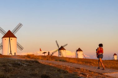 Toprak yolda yürüyen bir kadın altın saatinde geleneksel İspanyol yel değirmenlerini izliyor.