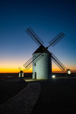 Renkli bir günbatımında tepede duran yel değirmenleri, ikonik La Mancha manzarasını temsil ediyor.