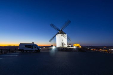 Campervan, alacakaranlıkta koyu mavi gökyüzünün altında tarihi İspanyol yel değirmenlerinin yakınına park edilmiş.