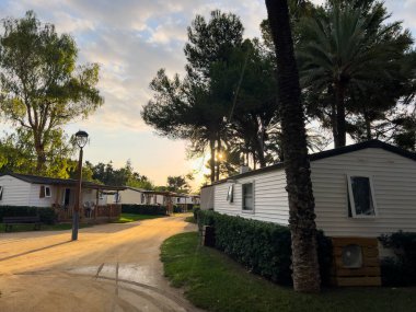 Gün batımında bir tatil parkında sıra sıra bungalovlar ve palmiye ağaçları, huzurlu bir akşamı gösteriyor.