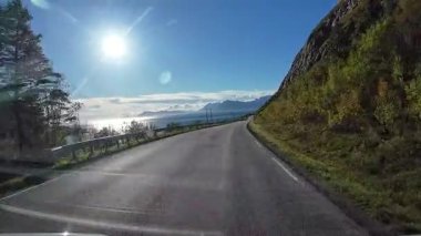 POV, Lofoten Adaları 'nda güneşli bir günde Norveç kıyı şeridi boyunca manzaralı bir yolda ilerliyor.
