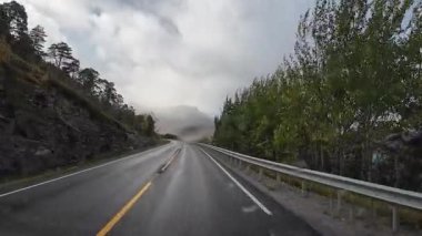 Norveç 'teki sisli bir İskandinav dağ manzarası boyunca ıslak asfalt bir yolda karavan sürüyor.