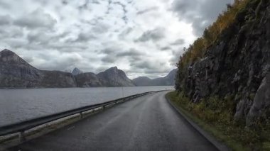POV, Norveç 'in kuzeyinde bulutlu bir günde bir fiyordun yanında dolambaçlı bir sahil yolunda ilerliyor.