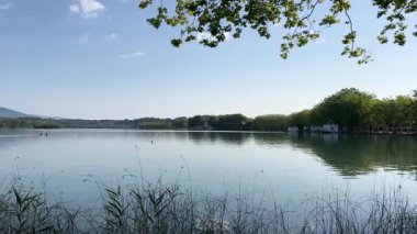 Girona 'daki sakin Banyoles Gölü' nün panoramik manzarası, güneşli bir günde insanların kürek çekmesi.