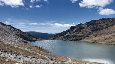 Bir grup yürüyüşçünün el değmemiş bir gölün yanında yürüdüğü göz kamaştırıcı bir dağ manzarası.
