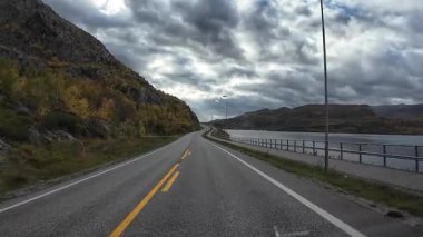 Kuzey Norveç 'te bir fiyort boyunca bulutlu bir sonbahar gününde manzaralı bir yolda araba sürmek