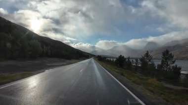 Islak bir yolda, güneşli ve bulutlu İskandinav kırsalında bir karavan yolculuğu.