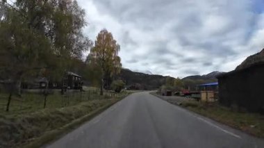 Bulutlu bir sonbahar gününde Norveç dağlarında kırsal bir yolda araba kullanan ilk kişi görülüyor.