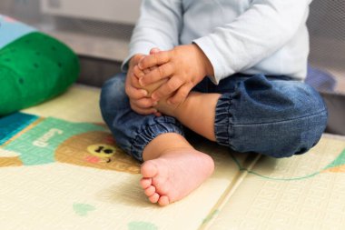 Renkli bir oyun hasırında oturan, ayakları keşfeden, kot pantolon ve açık renk gömlek giyen çocuk.