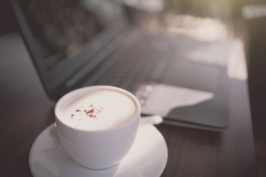 Beyaz kahve fincanı, bilgisayarın arka planında bulanık olan ahşap masanın üzerinde. Drama tonu.