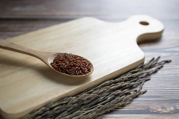 Red rice in wooden spoon on wooden cutting board and ears of rice on table.