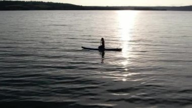  Yang kadınının silueti SUP tahtasının üzerinde duruyor ve gün batımında kürek çekiyor, suda dalgalanıyor. Havadan yan görüş. Güzel manzara. Fitness konsepti