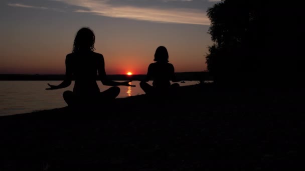 二人の柔軟な女の子がペアヨガの演習を行うシルエット サンセット屋外でビーチで一緒に水の近くにストレッチ スポーツトレーニング 精神調和のための心体操 物理的な健康 — ストック動画