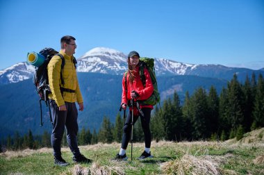 İki yürüyüşçü bir dağ sırtında mola veriyor. Sırt çantalı sportif çift, karlı dağların arka planında duruyorlar..