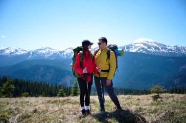 İki yürüyüşçü bir dağ sırtında mola veriyor. Sırt çantalı sportif çift, karlı dağların arka planında duruyorlar..
