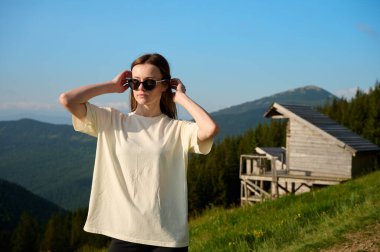 Kadın sıcak güneşin altında çimenli dağ patikalarında yürüyor, ahşap kulübeler ve arka planda uzak tepelerle. Barışçıl doğa seyahati ve doğa yaşam tarzı.