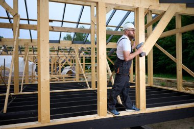 Ahşap çerçeve inşa ederken elektrikli tornavida kullanan inşaat işçisi. Koruyucu kasklı adam, eldiven. İnşaat, marangozluk, mühendislik ve inşaat süreci kavramı.