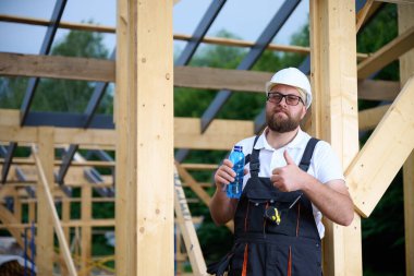 İnşaat işçisi ahşap ev iskeleti şantiyesinde mola verip su içiyor. İnşaatçı kaskı ve su şişesiyle dinleniyor..