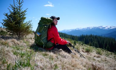 Çimenli dağ yamacında yürüyüş direğiyle dinlenen kadın yürüyüşçü. Kırmızı ceketli kadın, karlı tepelerin ve güneşli havanın tadını çıkarıyor. Açık hava seyahati ve macera yaşam tarzı.
