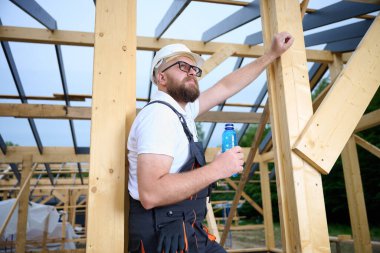 İnşaat işçisi ahşap ev iskeleti şantiyesinde mola verip su içiyor. İnşaatçı kaskı ve su şişesiyle dinleniyor..