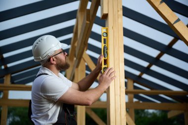 İnşaat işçisi, çerçeve alanındaki ahşap kirişin dikey hizasını kontrol etmek için ruh seviyesini kullanıyor. Kasketli ve güvenlik gözlüklü bir inşaat işçisi açık havada ahşap bir yapı üzerinde çalışıyor..