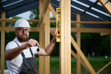 İnşaat işçisi anahtar tutarken ahşap kirişi kontrol etmek için ruh seviyesini kullanıyor. Kereste çerçeveli bina inşaatında çalışan kasklı ve tulumlu bir bina..