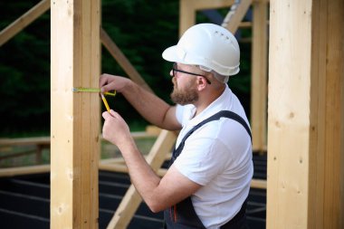 İnşaat işçisi inşaat iskeletinde tahta kirişi mezurayla ölçüyor. Kasklı bir bina, ahşap bina inşaatında çalışıyor..