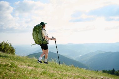 Bir kadın çimenli bir tepede büyük bir sırt çantası ve yürüyüş direkleri ile dikiliyor, uzak dağların manzarasının ve parlak bir gökyüzünün altındaki kır kulübesinin tadını çıkarıyor. Dışarıyı kucaklıyor..