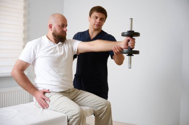 Fizyoterapist, erkek hastaya omuz rehabilitasyon egzersizinde yardımcı oluyor. Doktor gözetiminde fizyoterapi yaparken tıbbi kanepede oturan bir adam..