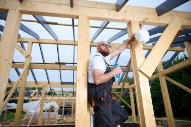 İnşaat işçisi ahşap ev iskeleti şantiyesinde mola verip su içiyor. İnşaatçı kaskı ve su şişesiyle dinleniyor..