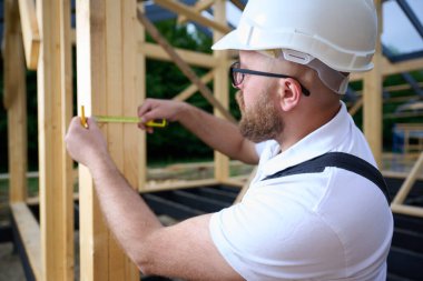 İnşaat işçisi inşaat iskeletinde tahta kirişi mezurayla ölçüyor. Kasklı bir bina, ahşap bina inşaatında çalışıyor..