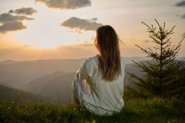 Kadın huzur içinde çimenli bir tepede oturur altın ışığın manzarayı yıkamasını seyreder. Yuvarlanan dağlar ve yemyeşil bir ağaç akşamları sakin bir atmosfer yaratır..