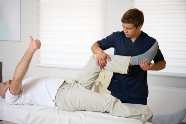 Modern tıp kliniğindeki rehabilitasyon seansında hastanın diz eklemlerini muayene eden fizyoterapist. Adam ortopedik tedavi görüyor ve fiziksel tedavi görüyor. Baş parmak işareti.